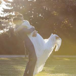 Le marié porte la mariée dans les bras et ils se font un calin dans les rayons du soleil avec des arbres en fond