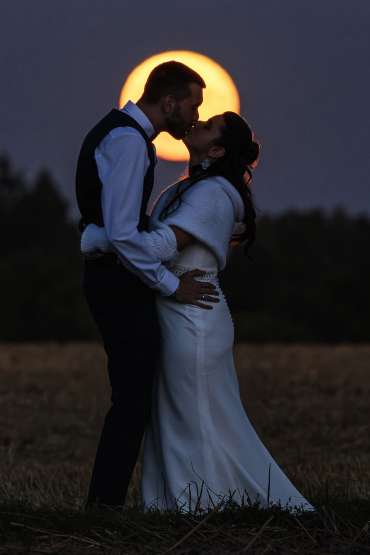 Couple de mariés très amoureux s'embrassant devant la pleine lune qui se lève au loin à la tombée de la nuit. Photo réalisée en août 2025 à 40 minutes de Maisons-Laffitte